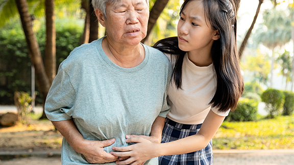 Elderly woman clenching waist with gastric discomfort as caregiver provides support.